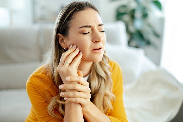 Upset caucasian woman in a yellow shirt sitting on a couch at home with eyes closed, holding her cheek in pain with a pained expression. Concept of toothache, dental pain, or jaw discomfort stock photo