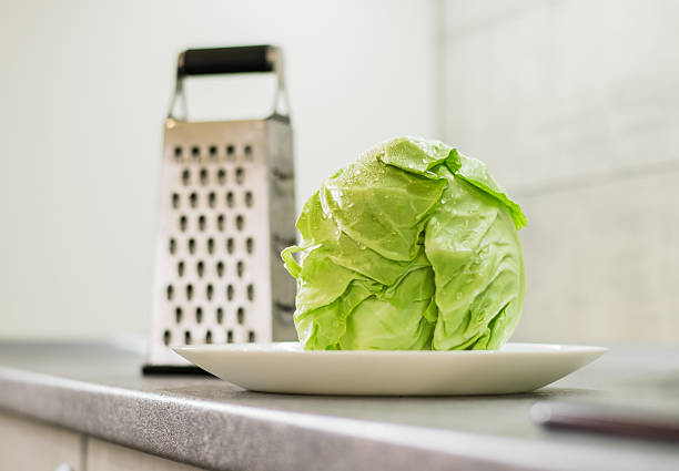 Cabbage with water droplets is placed on the kitchen countertop among metal tools. Light reflects off the grater, creating interesting shades on the surface of the cabbage. The green cabbage leaves add a natural look against the background of the neutral kitchen interior.