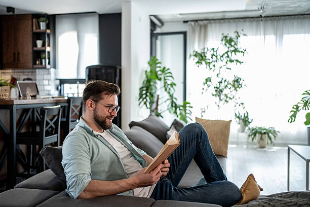 istock Young man relaxing on sofa reading a book in modern apartment 2209434907