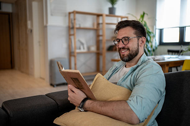 istock Smiling bearded man reading a book on a sofa at home 2209434929