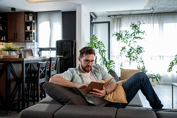 istock Young man reading a book on the sofa in his modern apartment 2209434951