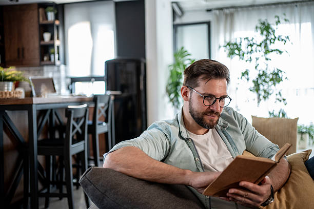 istock Young man reading a book on the sofa in his modern apartment 2209434989