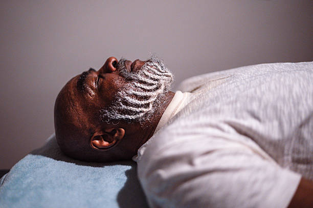 Side view of a healthy Black senior man with his beard braided laying with his eyes closed on a massage table during a mobility and stretching session at a luxury wellness center and spa.