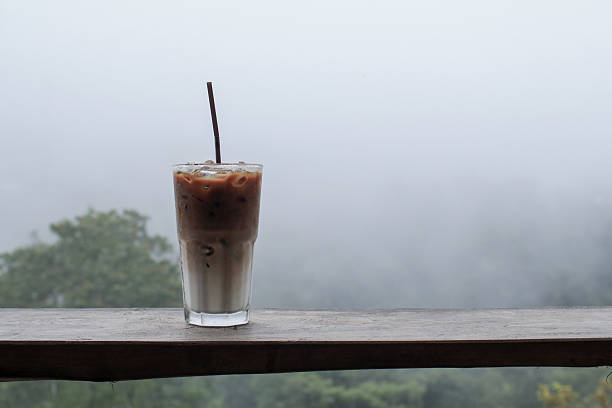 Iced coffee latte on wooden terrace after rain with misty fog on mountain background. Green Nature on holiday vacation.