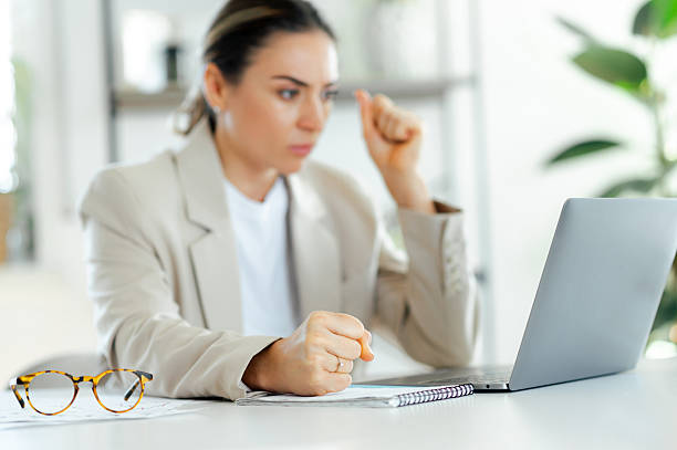 Defocused frustrated businesswoman in a beige blazer sits at a desk clenching her fist while looking at a laptop screen with a focused expression, looking dissatisfied and angry stock photo