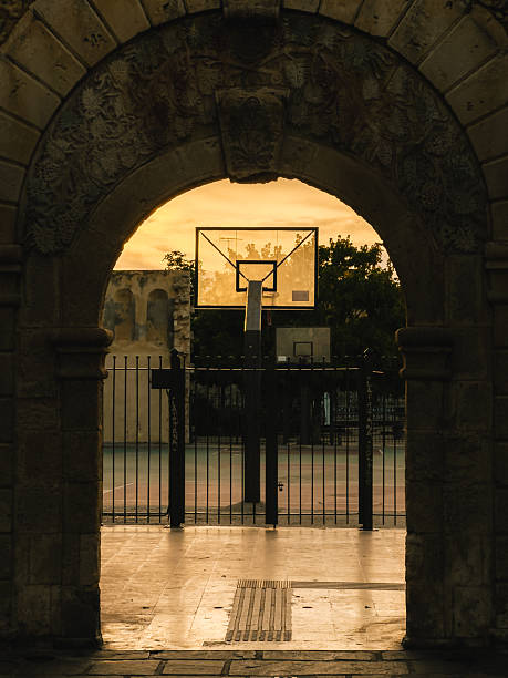 istock Basketball hoop standing behind gate near stone archway, sunset light casting long shadows across abandoned outdoor court, architectural contrast highlighting urban landscape. Basketball hoop, archway 2209462187