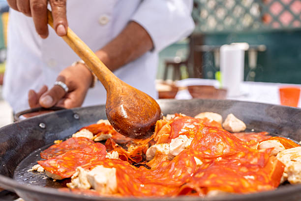 Chef using wooden spoon to stir chicken and chorizo while preparing paella outdoors on rooftop
