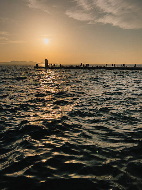Tourists silhouettes enjoying sunset at waterfront, with sun setting behind lighthouse and reflecting on sea creating golden waves. Silhouetted tourists watching sunset near lighthouse, golden hours