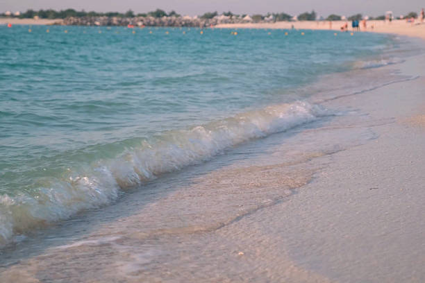 Gentle waves softly wash the sandy shore of a tranquil UAE beach, inviting relaxation and leisure. Distant figures enjoy the serene ambiance of the coastline in a peaceful setting
