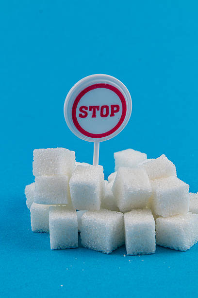 A pile of white sugar cubes is displayed against a bright blue background. A red and white stop sign is placed in the center, suggesting a warning about excessive sugar intake.