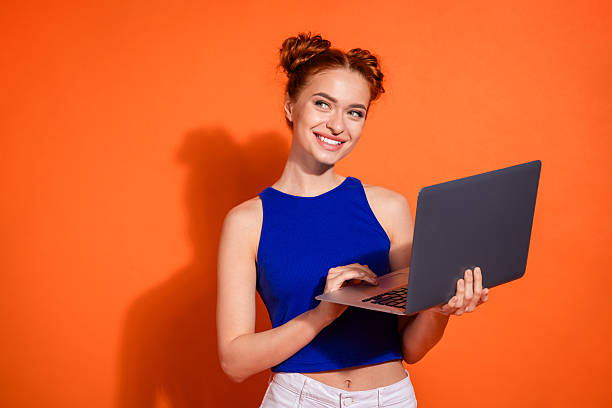 istock Young woman with a cheerful expression using a laptop against an orange background, wearing a stylish blue top 2209495265