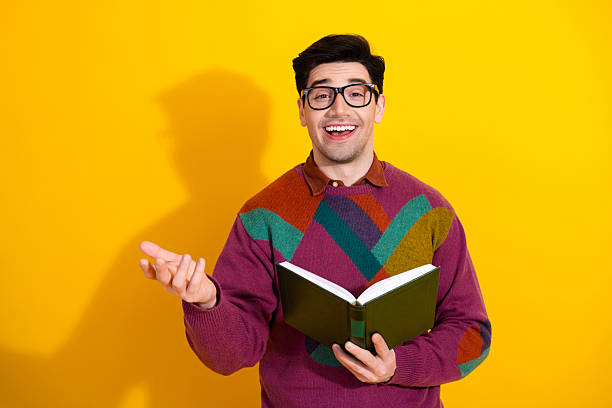 istock Young male with book gesturing confidently, wearing colorful pullover, standing against vibrant yellow background 2209498976