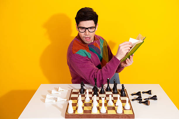 istock Young man in colorful sweater reading book while playing chess on vibrant yellow background, expressing creative and strategic thinking 2209499059