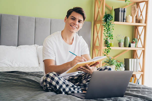 istock Young man in pajamas writing notes on bed, smiling in a comfortable well-lit bedroom, surrounded by plants and modern decor 2209504483