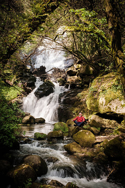 Man practicing yoga and meditating near waterfall in lush green forest