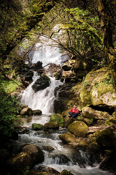 Man practicing yoga and meditating near waterfall in lush green forest
