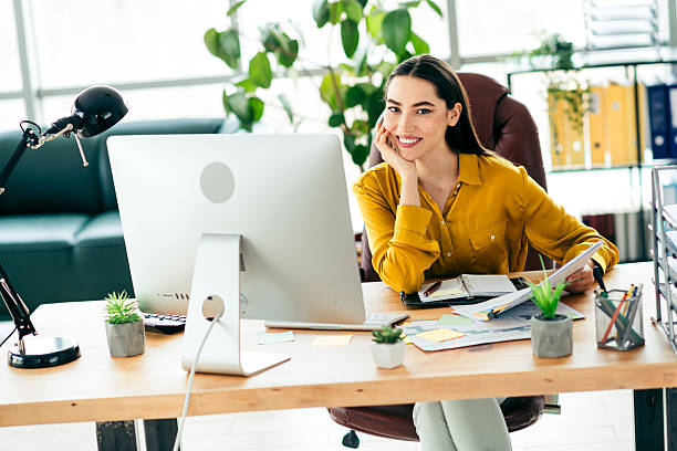Qualified professional woman smiling confidently at her desk in a modern office space surrounded by natural light and stylish decor Professional young lady sitting and working at her contemporary workspace with bright lighting. She exudes confidence while interacting within her business environment, portraying dedication and elegance. qualified report stock pictures, royalty-free photos & images