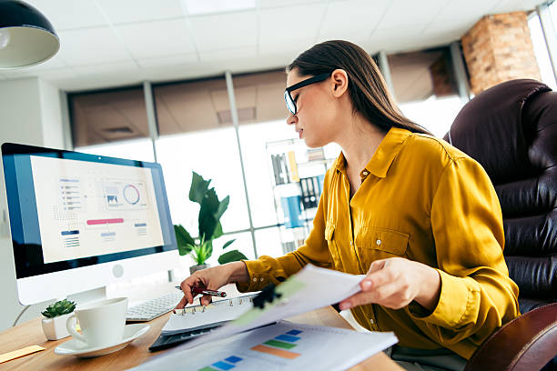 Qualified businesswoman working in a modern office environment analyzing financial documents and charts on her computer A professional woman in a yellow shirt working in an office, reviewing financial charts on a computer with documents in hand. Modern workplace atmosphere. qualified report stock pictures, royalty-free photos & images