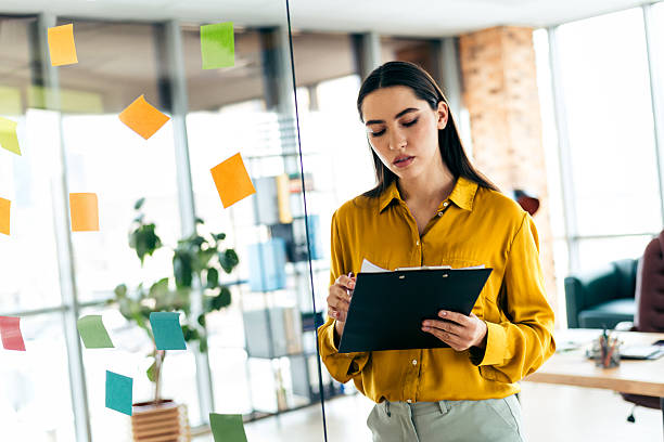 istock Young professional woman reviewing notes in a modern office setting with colorful sticky notes on a glass wall 2209507397