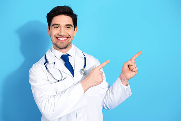 istock Young doctor in white coat smiles confidently and points towards a noteworthy direction against a bright blue background 2209507761