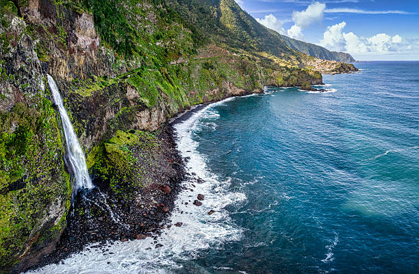 Aerial perspective revealing Bride's Veil waterfall plunging from rugged cliffside, tumbling toward azure Atlantic waters surrounding verdant Madeira Island landscape
