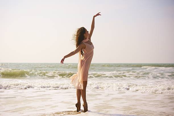 A romantic scene of a ballerina standing en pointe at the oceans edge, wearing a soft pink dress. Warm sunset light adds dreamy tones to the elegant moment in nature