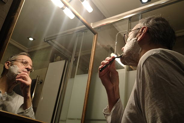 A shirtless man applies shaving cream with a razor in a tiled bathroom, seen in the mirror during a quiet grooming routine and personal care moment.