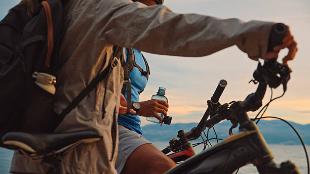 Close-up perspective of mid-adult Black male and Asian female cyclists taking water break during early morning ride, with ocean and mountain silhouettes creating dramatic backdrop.