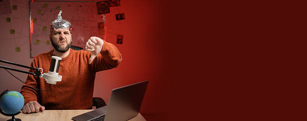 istock Bearded man in aluminum foil hat show thumb down dislike gesture reject refuse while Live streaming, presenter with microphone looking at camera. Conspiracy theory concept. Insanity. Banner. 2209528077