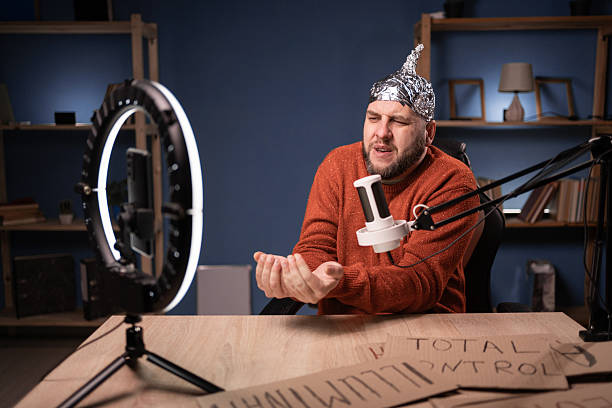 istock Paranoid man wearing foil hat recording video in home studio hope help, hands gesture. Sitting at desk with ring lamp and smartphone, sharing theories, creating unique content for social media 2209530874