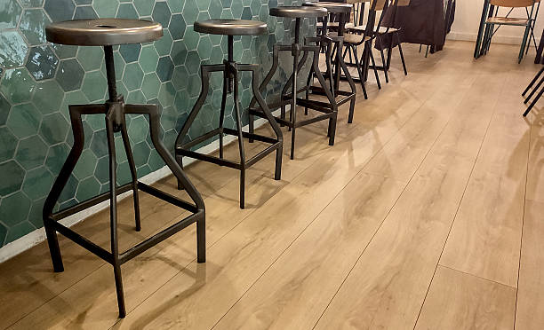 Industrial-style bar stools lined up against a green hexagonal tiled wall, with wooden flooring creating a warm, modern café or restaurant ambiance.