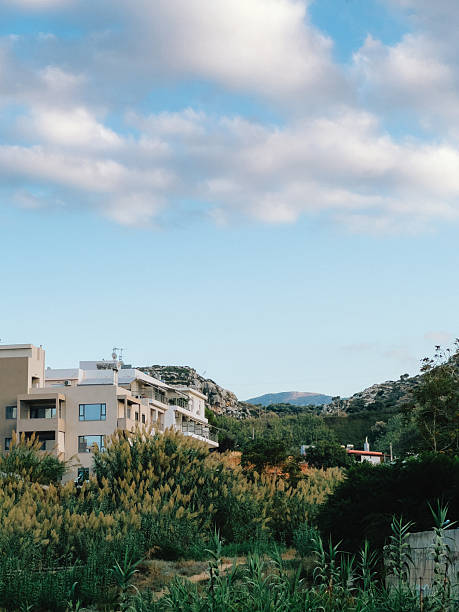istock Lush green vegetation partially obscures modern hotel against rolling hills under dynamic, cloud filled sky, creating tranquil and inviting scene. Verdant landscape, undulating terrain, cloudscape 2209532128