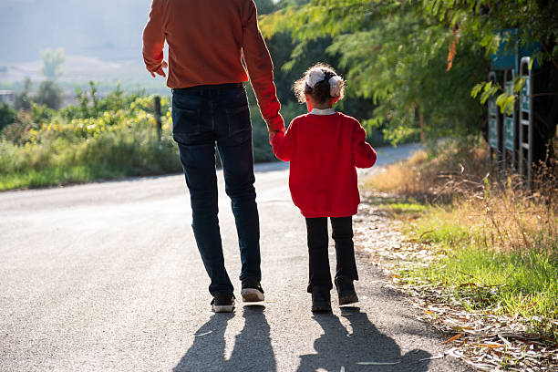 A father and daughter hold hands while strolling along a scenic path surrounded by greenery. The warmth of their bond is evident, showcasing family togetherness and outdoor enjoyment on a sunny day