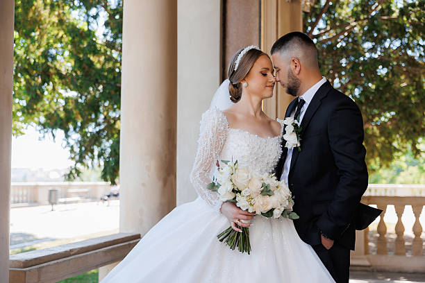 A bride and groom are standing on a balcony, with the bride holding a bouquet of flowers. Scene is romantic and happy, as the couple is embracing and looking into each other's eyes