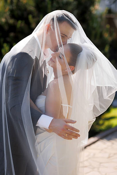 A bride and groom are hugging under a white veil. The bride is wearing a white dress and the groom is wearing a gray suit