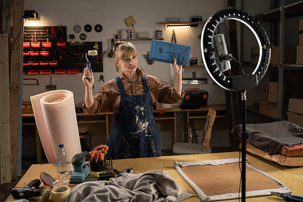 istock Middle aged craftswoman filming a tutorial about restoring furniture in her workshop using smartphone and ring lamp. She showing tray and paint roller 2209558273
