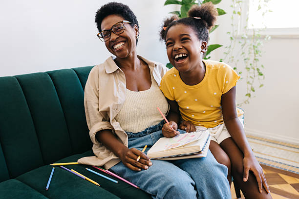 Happy young girl sitting with her mom o the couch, laughing and having fun as they engage in a colour drawing activity. Mother and daughter creating a fun, educational environment at home.
