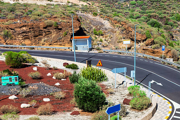 Winding mountain road curves through a rugged landscape with rocky cliffs and sparse greenery, featuring a small roadside booth. Warning signs and a vintage van add a touch of adventure