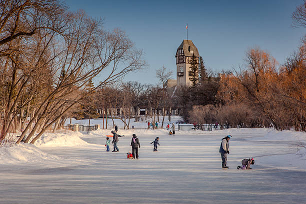 Winnipeg, February 7, 2009:  An outdoor scene capturing a lively winter day at Assiniboine Park.  Skaters glide accross the frozen pond.  The iconic pavilion stands proudly in the background, its historic architecture partially framed by bare trees.  A low winter sun casts a golden, warm light across the scene.