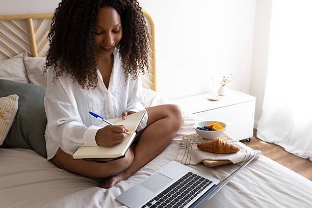 istock Young woman working from home taking notes in bed with laptop and breakfast 2209583807