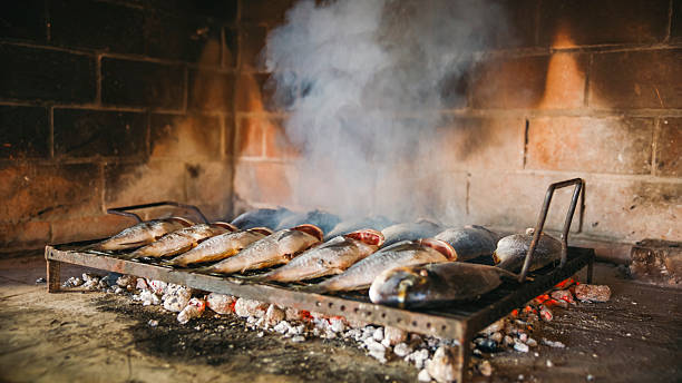 Fresh sea bream grilling on a metal rack over glowing charcoal in a rustic brick oven. Smoke swirls gently, enhancing the traditional cooking ambiance.
