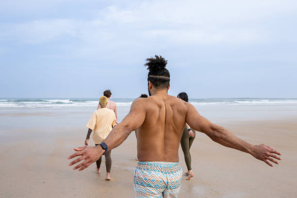 Rear view shot of a group of friends going for a sea dip at the beach together in Newton-by-the Sea, Northumberland, North East England. They are wearing active clothes and swimwear, walking towards the sea. Focus is on one man who is stretching his arms out behind him.