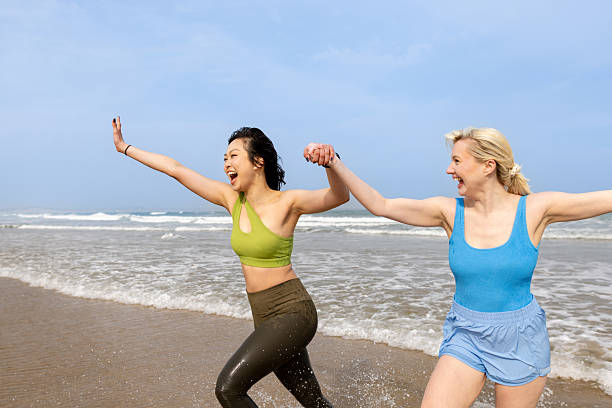 Two young women going for a sea dip at the beach together in Newton-by-the Sea, Northumberland, North East England. They are wearing active clothes and swimwear, having fun together outdoors. They're running away from the sea together, holding hands.