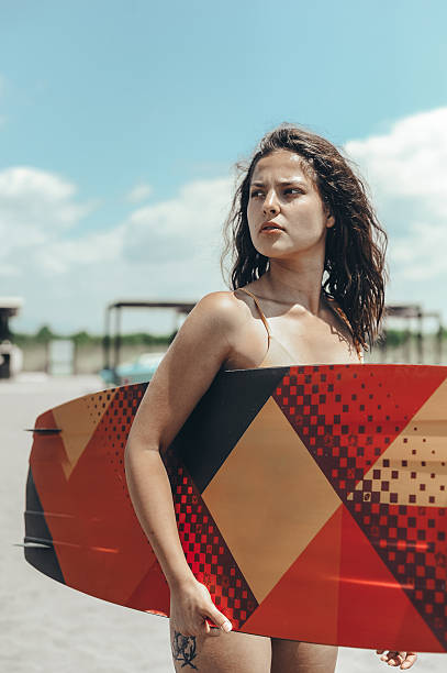 A young woman in a swimsuit holds a colorful surfboard while standing on a sunny beach with a bright blue sky in the background.