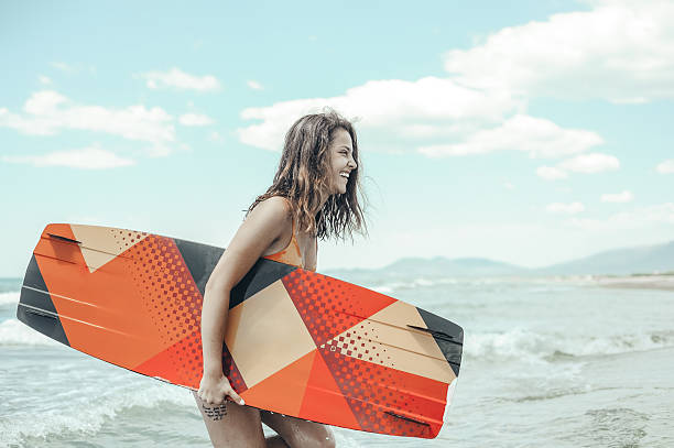 A young woman is walking along the beach carrying a vibrant, geometric-patterned surfboard with a smile on her face. The ocean waves and cloudy sky create a serene backdrop.