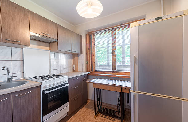 A cozy and well-organized kitchen space featuring modern wooden cabinetry, tiled backsplash, and a large window with blinds offering natural light