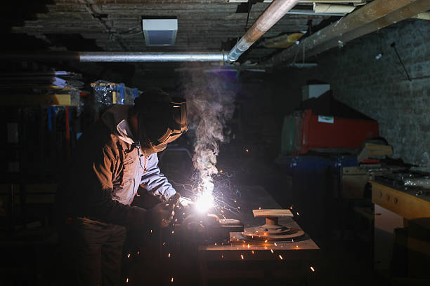 Close-up of a factory worker wearing protective gear and welding a piece of metal, with bright sparks and smoke visible. The background is dark, suggesting a low-light industrial environment. The image captures the intensity and precision of manual labor in a workshop setting.
