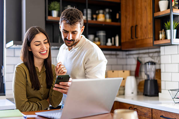 couple using laptop and smartphone, checking their home finances in the kitchen - huiseigenaar stockfoto's en -beelden