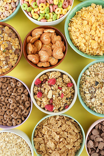 Full frame image of bowls of dry breakfast cereal and palmier biscuits, bran flakes, muesli, rolled porridge oats, chocolate flavoured crunchy wheat shells, chocolate and fruit flavoured O-shapes, corn flakes, granola, yellow background, elevated view