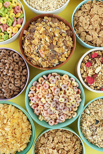 Full frame image of bowls of dry breakfast cereal, bran flakes, muesli, rolled porridge oats, chocolate flavoured crunchy wheat shells, chocolate and fruit flavoured O-shapes, corn flakes, granola, yellow background, elevated view
